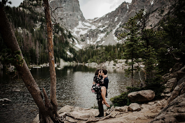 A young couple lean in for a kiss, while sitting on a fallen tree in a forest.